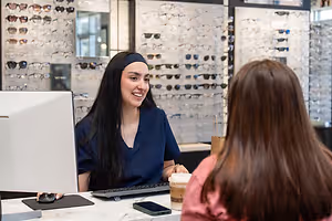 A woman in a blue shirt is sitting at a desk with a computer monitor and keyboard, smiling and looking at a woman in front of her who is holding a cup, while a monitor and a wall with sunglasses are behind her.