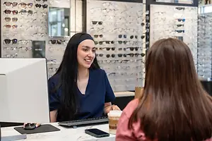 A woman in a blue shirt is sitting at a desk with a computer monitor and keyboard, smiling and looking at a woman in front of her who is holding a cup, while a monitor and a wall with sunglasses are behind her.