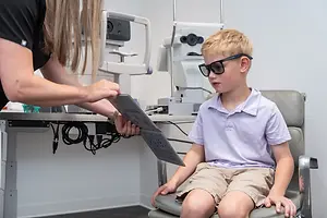 A boy is sitting in a chair at an optometrist's office, and a woman in a black shirt is examining him.