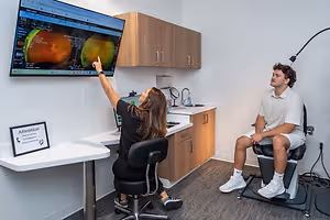 A woman points to an eye scan on a monitor while a man sits in a chair nearby in a medical office.