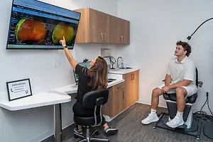 A woman points to an eye scan on a monitor while a man sits in a chair nearby in a medical office.