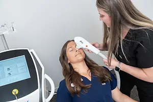 A woman is getting a facial treatment in a clinic by another woman who is wearing a black shirt and a watch.