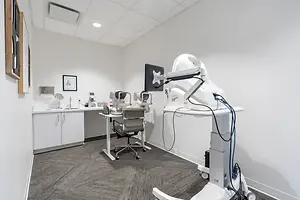 A medical equipment with a monitor is inside a room with a white wall and carpeted floor. There is a white desk with a chair and a monitor on top of it. On the left side, there is a cabinet with a sink and a picture frame mounted on the wall. There is also a picture frame hanging on the wall. The ceiling has a white square-shaped design.
