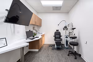 A white room with a desk, monitor, keyboard, chair, and medical equipment on the right side.