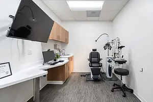 A white room with a desk, monitor, keyboard, chair, and medical equipment on the right side.