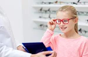 A young girl is trying on a pair of red glasses in a store with a man wearing a white coat holding a blue box with a pen inside