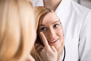 A smiling woman with a contact lens in her eye is being checked by a doctor in a white lab coat