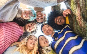 A group of people are smiling and posing for a photo, with one person wearing a gray sweater and another person wearing a pink sweater. They are all wearing winter clothes and appear to be standing in a circle. The person with blue hair is wearing a yellow shirt and has a big smile on his face. The person with a hat is wearing a blue and white striped shirt.