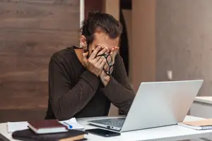 A man sitting at a desk with a laptop and books covering his face with his hands.