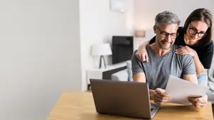 A couple is working on a laptop while sitting at a desk, the man is reading a paper and smiling, and the woman is looking at him while leaning on him. Behind them is a white wall with a lamp on the desk, an air conditioner on the wall, and a TV on the wall.