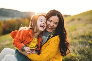 A mother and daughter are sitting on the grass, smiling and looking at each other, surrounded by grass and trees
