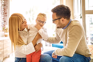 A family is sitting in a room with a smiling child in the middle. The woman on the left is holding the child, while the man on the right is looking at the child with a smile. Behind them is a window with a view of the outside.