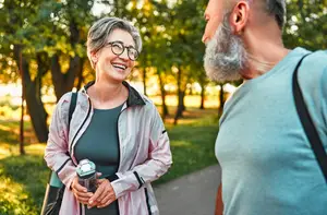 An adult woman with glasses is holding a bottle and smiling while walking with a man in a park with trees and grass.