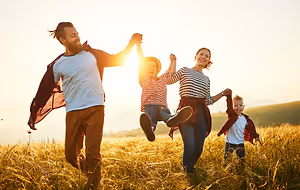 A family is jumping in a grassy field, with a man holding a young girl's hand, a woman holding a boy's hand, and a young girl jumping in the air with a smile on her face.