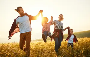A family is jumping in a grassy field, with a man holding a young girl's hand, a woman holding a boy's hand, and a young girl jumping in the air with a smile on her face.