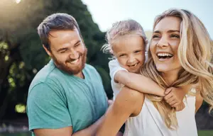 A family of three smiling and looking joyful in a park, the woman holding a child and the man standing beside them, both with smiles on their faces