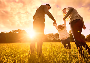 Three people are standing in a grassy field with the sun setting in the background, and they are holding hands, two of them are adults, and one is a child. The child is jumping and holding the hands of the adults. The adults are looking at the child, and the child is looking at the adults. The field is surrounded by trees and plants. The sky is bright and clear. The image has a blurry effect.