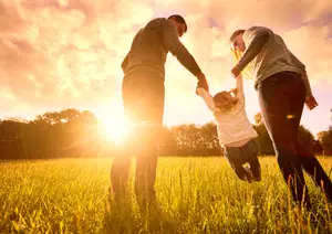 Three people are standing in a grassy field with the sun setting in the background, and they are holding hands, two of them are adults, and one is a child. The child is jumping and holding the hands of the adults. The adults are looking at the child, and the child is looking at the adults. The field is surrounded by trees and plants. The sky is bright and clear. The image has a blurry effect.