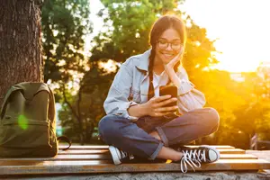 Young woman sitting on a bench in a park using a smartphone, with a backpack nearby, during a sunny day.
