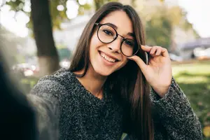 A woman with glasses and a sweater poses for a selfie in a park with trees and grass in the background.
