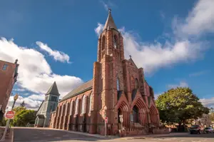 A large church with brick walls and a steeple is located on a city street with trees and a stop sign.