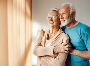 An elderly couple smiling and hugging each other in a room with sunlight coming through the window