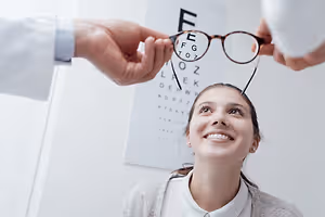 A smiling woman in a white shirt is trying on glasses in front of an eye chart, with a doctor holding the glasses behind her head.