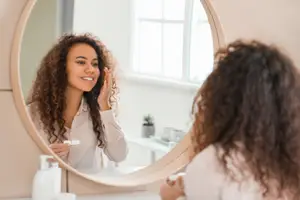 A woman with curly hair is brushing her teeth while looking in a mirror.