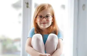 A young girl with red hair wearing glasses and a blue shirt is sitting in front of a window and smiling