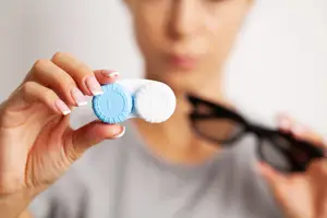 A woman holds a contact lens case in her hands, with her other hand holding a pair of glasses