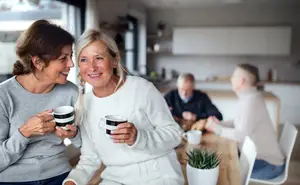 An older woman in a white sweater holds a coffee cup and laughs with another older woman in a gray sweater.