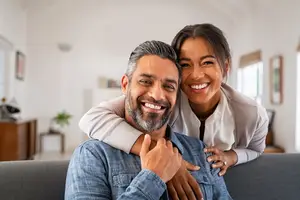 A man and woman are sitting on a couch in a living room, smiling and posing for a photo.