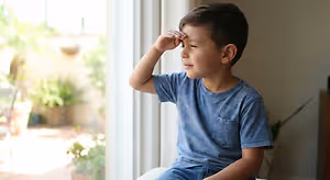 A young boy sitting by a window, shading his eyes from the sun with his hand.