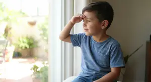 A young boy sitting by a window, shading his eyes from the sun with his hand.