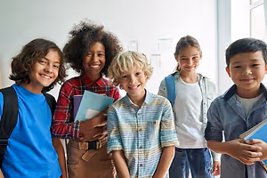 A group of smiling children standing in a classroom with some carrying books and backpacks
