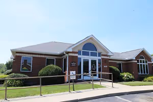 A brick building with glass windows and a glass door in front of a lawn with plants and bushes, a metal railing on the side, and a parking lot in front under a blue sky.