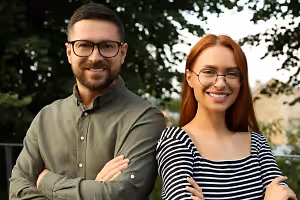 A man and a woman standing together in a park, both wearing glasses and smiling