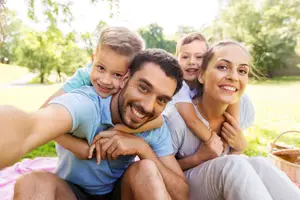 A happy family of four, a man, a woman, and two kids, are sitting on a grassy field in a park with a basket, looking at the camera and smiling.