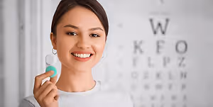 A woman is holding a green toothbrush in her hand and smiling at the camera. She is standing in front of a wall with a black border and a white background that has letters on it. The letters are in the order of W, K, F, O, L, P, Z, D, R, E, L, N, B, W, M, H, R, P, A, E, C, E, I, U, O, E, A, K, F, O, R, M, A, T, I, O, N. The woman is wearing a white long-sleeve shirt and has brown hair.
