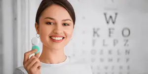 A woman is holding a green toothbrush in her hand and smiling at the camera. She is standing in front of a wall with a black border and a white background that has letters on it. The letters are in the order of W, K, F, O, L, P, Z, D, R, E, L, N, B, W, M, H, R, P, A, E, C, E, I, U, O, E, A, K, F, O, R, M, A, T, I, O, N. The woman is wearing a white long-sleeve shirt and has brown hair.