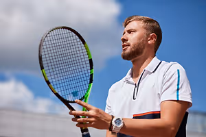 A man with a beard is holding a tennis racket and is looking up at the sky, possibly on a tennis court.