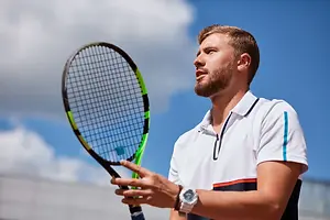A man with a beard is holding a tennis racket and is looking up at the sky, possibly on a tennis court.