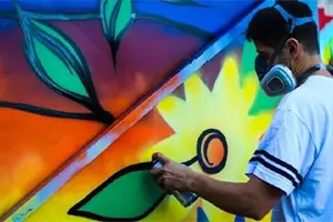 A man is spray painting a colorful mural on a wall with a mask on