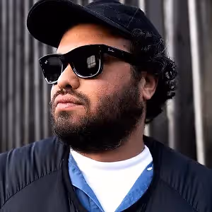 A man with a beard wearing a black hat and sunglasses is standing outside in front of a wall.