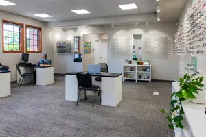 A room with white desks, chairs, a monitor, and a potted plant. A person is sitting on a chair and working on the desk. Another person is sitting in the doorway. The room has two windows and several lamps on the ceiling.