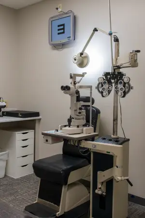 An ophthalmoscope machine and a black leather chair are in a room with a monitor and a desk with drawers