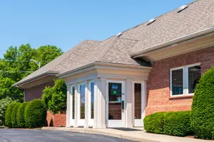 An image of a brick building with a gray roof and white trim and windows with a driveway and bushes in front
