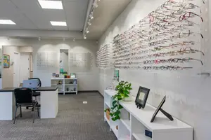 Interior view of a modern office with white walls, gray carpet, and various office equipment, including a desk, chairs, and a shelf displaying eyeglasses, with ceiling lights and a framed picture on the wall.
