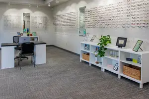A clean and organized office space with white furniture, a carpeted floor, and shelves displaying glasses and other items. The room has mirrors, a desk with a laptop, and a plant.