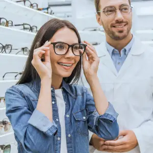 A woman trying on glasses in a store with an optician in the background